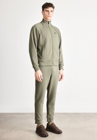 Φόρμα σε ελαιί πράσινο χρώμα από απαλό ύφασμα, με ψηλό γιακά, κλείσιμο με φερμουάρ και πλαϊνές τσέπες. Διαθέτει αντίθετες λευκές ρίγες κατά μήκος των πλαϊνών.