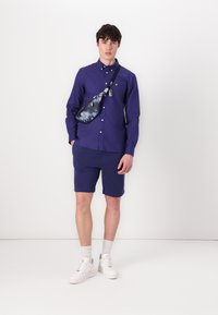 Camisa morada de manga larga con cuello abotonado, combinada con pantalones cortos a juego. Zapatillas blancas y un bolso cruzado de patrón completan el look.