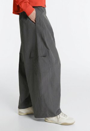 Person iført brede grå cargobukser, hvide sneakers og en rød langærmet top med den ene hånd i lommen.