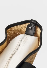 Geflochtene Strohtasche mit einem schwarzen Lederbesatz, die ein weißes Canvas-Reißverschlusspäckchen im Inneren hat. Die Tasche hat eine glatte Textur und eine strukturierte Form.