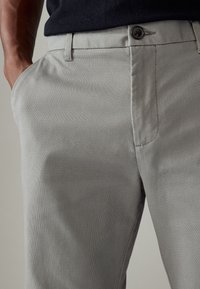 Pantalon gris clair en tissu texturé ; dispose de poches latérales inclinées, d'une fermeture à bouton et d'un design à plat à l'avant.