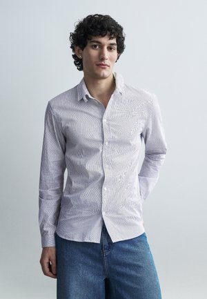 Noor mees lokkis juustega, seljas hele triibuline nööpidega särk ja sinised teksad, seisab ühevärvilise halli tausta ees.