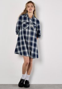 Niebieska i biała sukienka w kratę z guzikami na przodzie, dwoma kieszeniami na piersi, podwiniętymi rękawami i luźnym krojem. W zestawie z czarnymi, masywnymi butami i białymi skarpetkami.