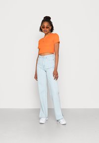 T-shirt curta laranja com bainha decorativa, jeans de cintura alta listrados em azul claro na vertical, e ténis brancos. Óculos de sol transparentes completam o visual.