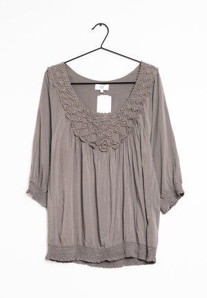 Blusa grigia leggera con maniche a 3/4, caratterizzata da un colletto in crochet testurizzato e un orlo arricciato, appesa a un gancio nero su uno sfondo bianco.