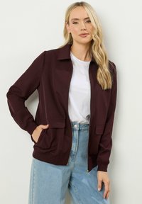 Bordeaux zip-up jack met twee voorzakken, geribde manchetten en een kraagnek, gecombineerd met een wit T-shirt en lichtblauwe jeans.