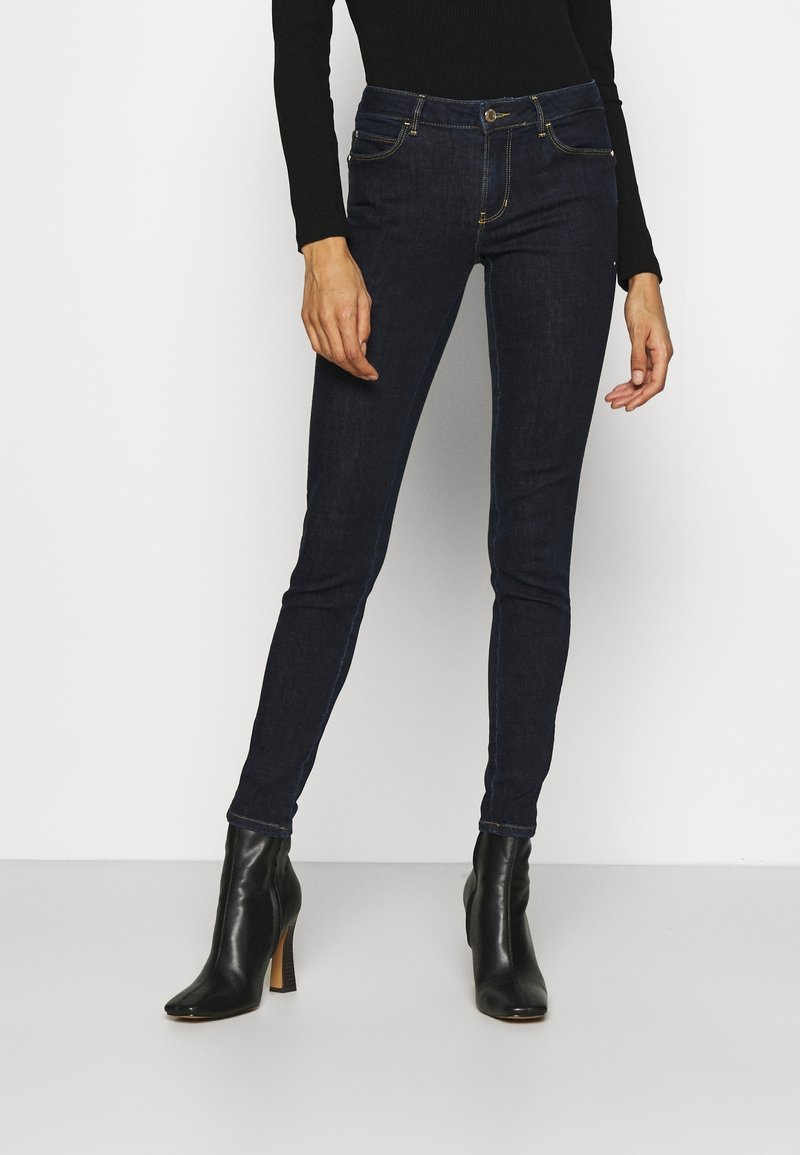 Mörkblå skinny jeans i denim med en slät yta, femfickorsdesign och guldstickningar. Bärs med svarta klackstövlar.