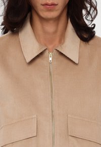Personne portant une veste beige en velours côtelé avec une fermeture éclair à l'avant et de grandes poches à rabat, vue de la moitié de la poitrine jusqu'au cou.