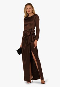 Długa, brązowa satynowa sukienka z długimi rękawami, bocznym rozcięciem i kokardą w talii. Noszona z sandałami na wysokim obcasie oraz małą torebką.
