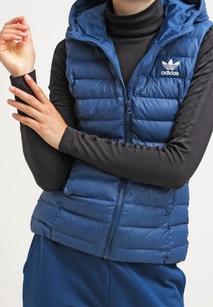 Osoba z założonymi na piersi rękami, ubrana w niebieską pikowaną kamizelkę Adidas z kapturem na czarnej bluzce z długim rękawem i niebieskich spodniach.