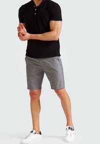 Mies joka on pukeutunut mustaan polo-paitaan, harmaisiin kuvioituihin shortsseihin ja valkoisiin lenkkareihin, seisoo kädet ristissä edessä.