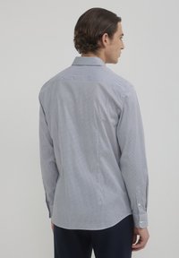 Homme portant une chemise à manches longues à motifs gris clair et un pantalon sombre, vu de dos contre un fond uni clair.