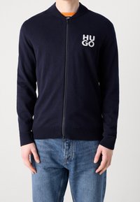 Navy zip-up cardigan i mjukt stickat tyg, med rundad krage, ribbade muddar och en vit "HUGO"-logotyp på bröstet.