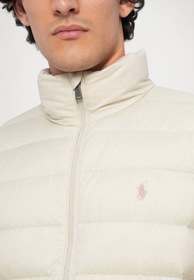 Giacca imbottita beige con colletto alto, zip frontale e un piccolo logo ricamato sul petto. Texture liscia e materiale leggero.