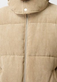 Beige corduroy puffer jacket featuring a high collar, vertical ribbed texture, and silver zipper down the front. Accompanied by snap closures.