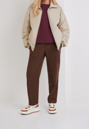 Persona con chaqueta acolchada beige, camiseta burdeos, pantalones marrones y zapatillas blancas con detalles marrones, de pie frente a un fondo claro y liso.