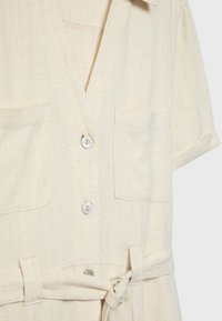 Chemise beige clair à manches courtes boutonnée, avec deux poches poitrine et une ceinture en tissu nouée à la taille.