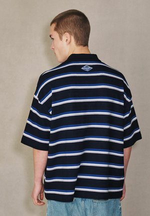 Jeune homme aux cheveux courts portant un polo bleu marine ample avec des rayures horizontales blanches et bleues, vu de dos sur un fond uni.