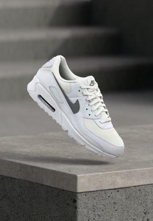 Pantof sport alb Nike Air Max cu logo Swoosh gri plutind deasupra unei margini de beton, prezentând detalii laterale și frontale.