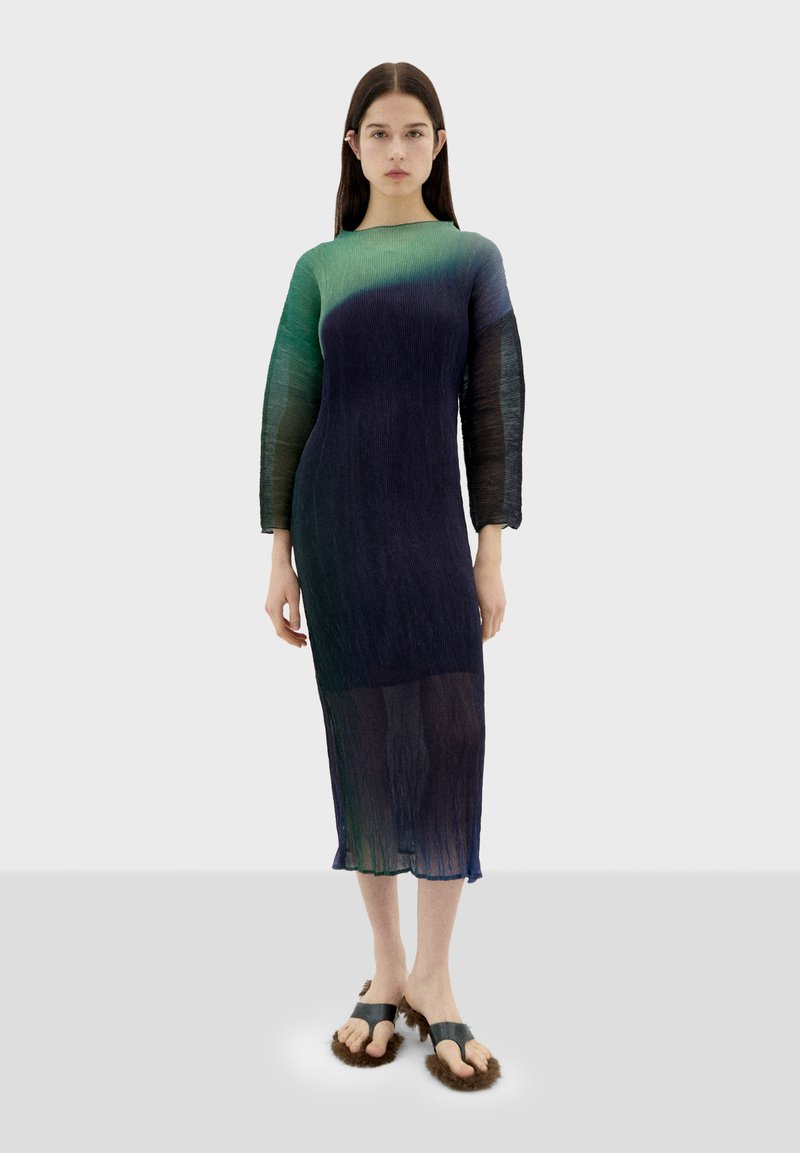 Fließendes, durchscheinendes Kleid mit langen Ärmeln, das am Ausschnitt in ein Grün übergeht und am Saum dunkelblau wird. Getragen mit flauschigen Sandalen.