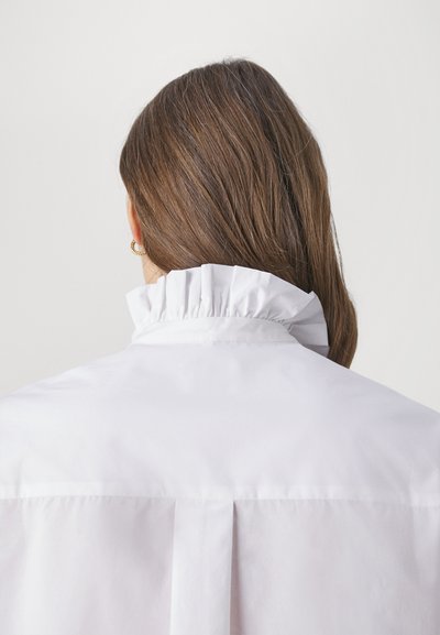 Frau mit langen braunen Haaren, die ein weißes Hemd mit hohem, voluminösem Kragen trägt, von hinten vor einem schlichten hellen Hintergrund dargestellt.