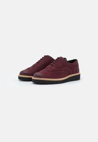 Une paire de chaussures brogues en suede bordeaux avec des détails perforés, des semelles intermédiaires marron clair et des semelles extérieures en caoutchouc noir sur un fond blanc.