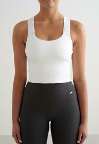 Valkoinen ribattu aktiivivaatteiden crop-top, jossa on säädettävät hihnat, yhdistettynä korkeavyötäröisiin mustiin ribattuihin legginsseihin, joissa on pieni metallinen logo.