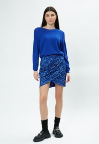 Blauer Strickpullover mit strukturierten Schultern, kombiniert mit einem hoch-niedrig geschnittenen blauen Rock, der ein gemustertes Design und Drapierungsdetails aufweist. Schwarze klobige Schuhe.