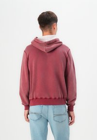Sudadera con capucha color burdeos, con puños y dobladillo acanalados, de textura suave y diseño en la parte posterior. Combinada con jeans de color azul claro con costuras visibles.