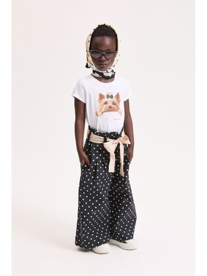 Bambino che indossa una maglietta bianca con stampa di cane, pantaloni neri a gamba larga a pois con cintura beige a fiocco, sciarpa a pois e occhiali da sole scuri.