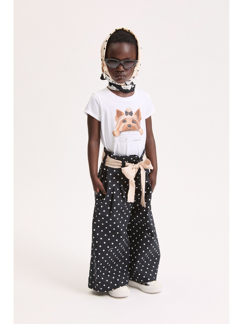 Παιδί που φοράει λευκό μπλουζάκι με στάμπα σκύλου, μαύρο παντελόνι με φαρδιά πόδια και πουά σχέδιο, μπεζ ζώνη με φιόγκο, μαντήλι με πουά και σκούρα γυαλιά ηλίου.