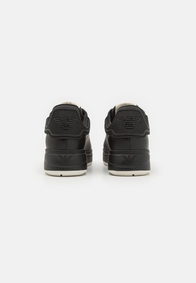 Black sneakers with textured rear panel, smooth upper, and contrasting white sole. Features logo detail on back and padded collar.