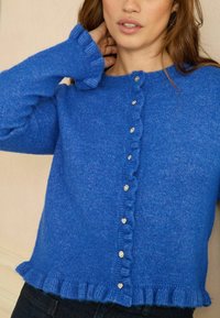 Nő élénk kék kötött kardigánban, fodros szegélyekkel és apró, szív alakú kristálygombokkal az elején.