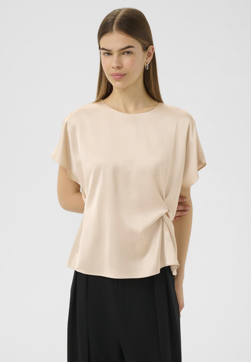 Haut beige à manches courtes en tissu lisse, avec un col rond et un détail noué sur le côté, associé à un pantalon noir.