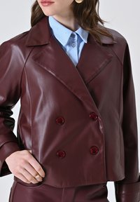 Vrouw in een bordeauxrode leren dubbelbreasted jas over een lichtblauw overhemd, met haar hand op haar heup en een gouden ring om haar vinger.