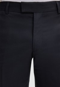 Dunkelblaue, maßgeschneiderte Hose mit strukturierter Oberfläche, vorne glatt, mit Gürtelschlaufen und scharfen Bügelfalten entlang der Beine.