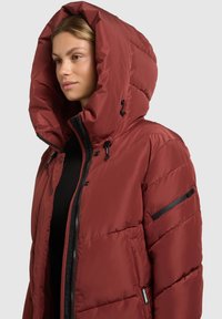 Giacca a piumino rossa con un ampio cappuccio, dotata di una cerniera frontale e tasche laterali. Il tessuto è liscio e leggermente lucido, con dettagli trapuntati.