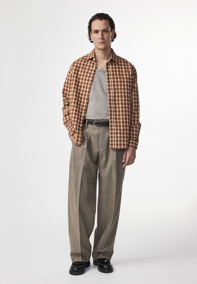 Braun-weiße karierte Button-Up-Bluse über einem grauen Tanktop, kombiniert mit hellbraunen gefalzten Hosen und schwarzen Schuhen.