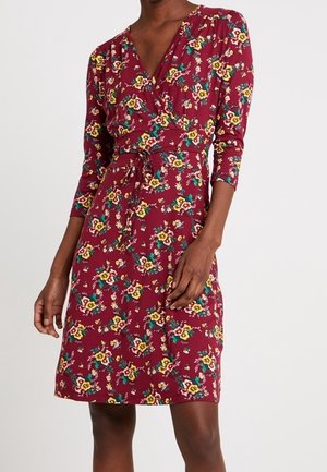 Femme portant une robe portefeuille mi-longue à manches longues, fond rouge avec imprimé floral jaune, nouée à la taille avec une ceinture en tissu assorti.