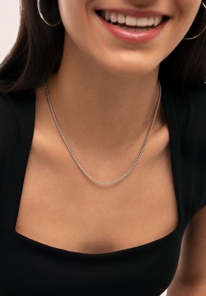 Collier en chaîne argentée reposant sur une clavicule nue, avec un design de maillons décorés, porté sur un haut noir à encolure carrée.