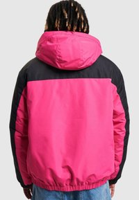 Rosa og svart hettejakke med elastiske mansjetter og midjebånd. Har et stort fargeblokk-design og glatt tekstur.