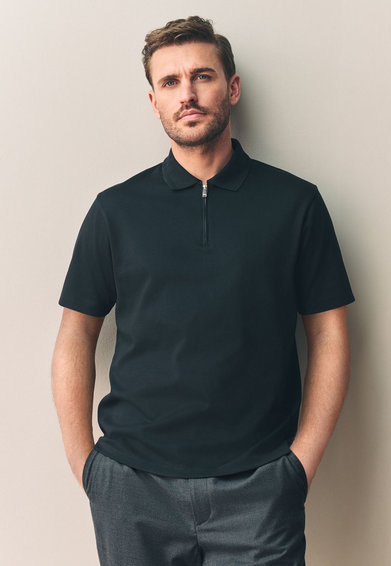 Zwart poloshirt met korte mouwen en een ritskraag, gemaakt van gladde stof. Draagt grijze broek, handen in de zakken.