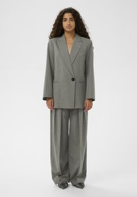 Femeie cu păr lung și creț, purtând un costum oversized cu dungi gri, cu pantaloni evazați și pantofi cu vârf ascuțit, pe un fundal simplu.