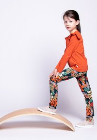 Pomarańczowa bluzka z długim rękawem z falbanką, wielokolorowe legginsy w kwiaty oraz białe buty wsuwane, pozujące na zakrzywionej drewnianej powierzchni.