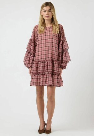 Femme portant une robe ample à carreaux roses et bruns avec des manches et un ourlet à volants étagés, associée à des talons pointus marron, debout devant un fond blanc.