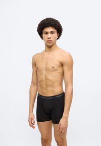 Μαύρα βαμβακερά ανδρικά μποξεράκια με φαρδιά ελαστική μέση που φέρει το λογότυπο "TOMMY HILFIGER" σε αντιθετικά χρώματα. Το μοντέλο στέκεται χωρίς μπλούζα.