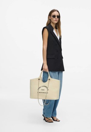 Femme portant un blazer noir sans manches, jean bleu, sandales noires à talons, lunettes de soleil, tenant un grand sac cabas beige Karl Lagerfeld avec une bandoulière chaîne.