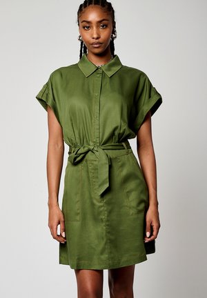 Robe chemise vert olive avec des manches courtes enroulées, un col, une ceinture à nouer à la taille et des poches avant. Tissu lisse avec des coutures visibles.