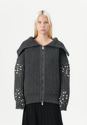Femme aux cheveux blonds raides portant un pull zippé gris foncé à côtes avec un large col et des manches ornées, associé à une jupe noire.