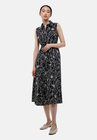 Robe noire avec motif floral blanc, sans manches, col rabattu, boutons sur le devant et jupe fluide. Chaussures plates beiges.